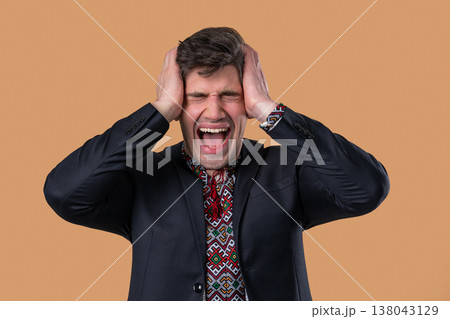 Stressed ukrainian businessman grabs head in panic, screaming on beige studio. Stressed ukrainian businessman grabs head in panic, screaming on beige studio. 138043129