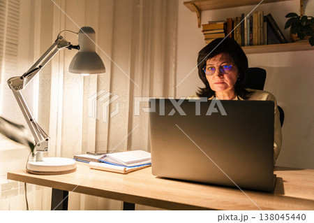 Woman focusing on her laptop screen while working remotely from home in evening, illuminating desk area, working late at night Woman focusing on her laptop screen while working remotely from home in evening, illuminating desk area, working late at night 138045440