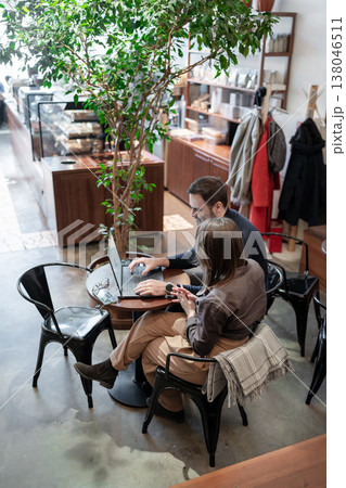 Business meeting at coffee shop, partners discussing project strategy across cups and plants. Business meeting, coffee shop, strategy, partners, teamwork. Business meeting at coffee shop, partners discussing project strategy across cups and plants. Business meeting, coffee shop, strategy, partners, teamwork. 138046511