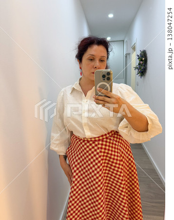 Woman taking mirror selfie in white short sleeve blouse and red gingham midi skirt at home. Outfit coordination, wardrobe styling, everyday elegance, personal fashion documentation, feminine clothing 138047254