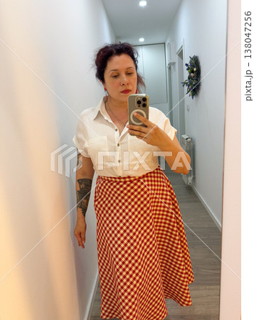Woman taking mirror selfie in white short sleeve blouse and red gingham midi skirt at home. Outfit coordination, wardrobe styling, everyday elegance, personal fashion documentation, feminine clothing 138047256