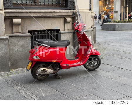 Red motor scooter parked beside stone building on paved city sidewalk near a commercial pedestrian area. Urban mobility, compact transportation, independent commuting, street parking, everyday 138047267