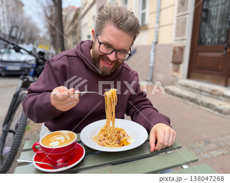 Bearded man in burgundy hoodie eating spaghetti and drinking latte at outdoor cafe table on a quiet city street. Casual urban dining, weekend brunch culture, neighborhood hospitality, everyday social 138047268