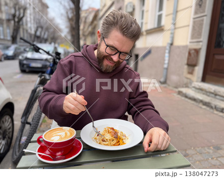 Bearded man twirling spaghetti with fork while sitting at an outdoor cafe table beside a red latte cup. Street dining ritual, relaxed meal break, urban cafe culture, approachable everyday lifestyle Bearded man twirling spaghetti with fork while sitting at an outdoor cafe table beside a red latte cup. Street dining ritual, relaxed meal break, urban cafe culture, approachable everyday lifestyle 138047273