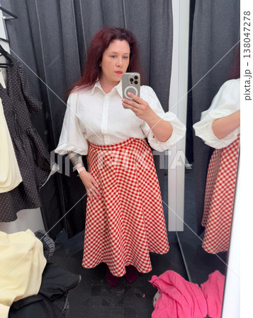 Woman taking mirror selfie in fitting room wearing white blouse with red gingham midi skirt. Outfit coordination, boutique styling, garment comparison, wardrobe planning, personal fashion selection Woman taking mirror selfie in fitting room wearing white blouse with red gingham midi skirt. Outfit coordination, boutique styling, garment comparison, wardrobe planning, personal fashion selection 138047278