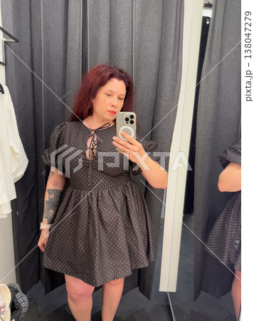 Woman taking mirror selfie in fitting room wearing black mini dress with dotted fabric. Dress fitting, fashion purchase, boutique browsing, outfit evaluation, personal style experimentation in retail 138047279