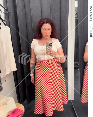 Woman taking mirror selfie in fitting room wearing cream knit top and red gingham midi skirt. Everyday fashion styling, garment fitting, retail decision making, personal wardrobe selection 138047281