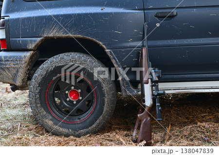 Hunting rifle leaning against a muddy off-road truck wheel after a rural field hunt 138047839
