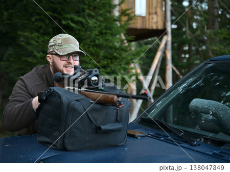 Hunter aiming a scoped rifle from vehicle hood with shooting rest near wooden hunting stand in forest 138047849