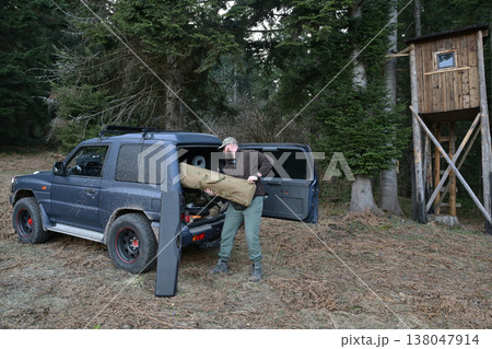 Man loading hunting gear into a muddy SUV near a wooden deer stand in a forest 138047914