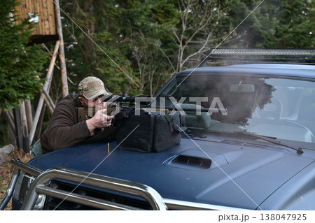 Hunter taking aim with a scoped rifle from a pickup truck hood in a forested setting 138047925