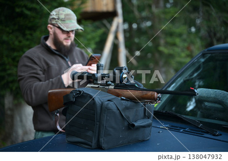 Hunter preparing scoped rifle on truck hood while checking phone near wooded hunting stand 138047932