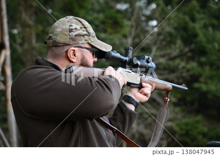 Hunter aiming a scoped rifle while wearing a camouflage cap in a forest setting 138047945