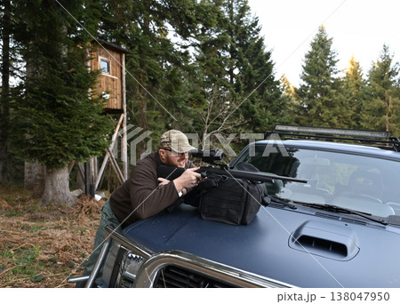 Hunter aiming scoped rifle from pickup truck hood near a wooden hunting blind in evergreen forest 138047950
