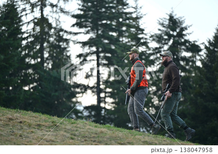 Two hunters walking through forest hillside carrying rifles and wearing hunting gear and orange vest Two hunters walking through forest hillside carrying rifles and wearing hunting gear and orange vest 138047958