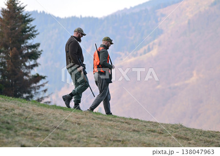 Two hunters walking down a mountain slope carrying rifles during autumn hunting trip Two hunters walking down a mountain slope carrying rifles during autumn hunting trip 138047963