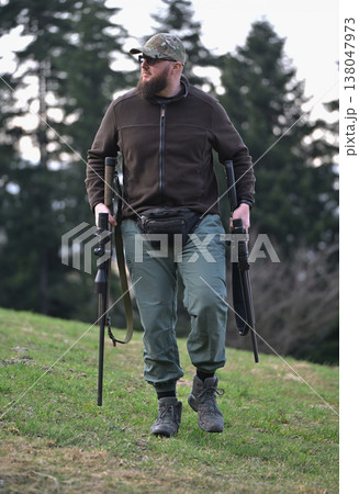 Bearded man carrying rifles walking on grassy hill in outdoor hunting gear and camouflage cap 138047973