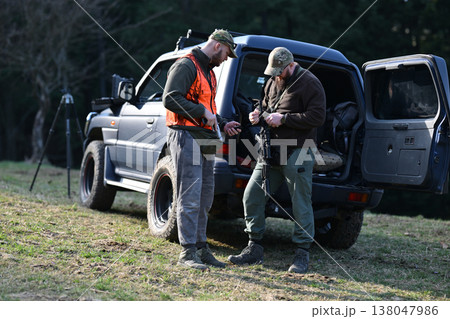 Two hunters preparing rifles by an off-road vehicle in a forest clearing before a hunt 138047986