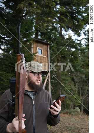 Hunter with rifle and smartphone near a wooden hunting blind in a forested outdoor setting Hunter with rifle and smartphone near a wooden hunting blind in a forested outdoor setting 138048000