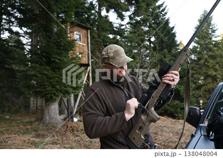 Hunter adjusting scoped rifle near a wooden deer stand in dense evergreen woods 138048004