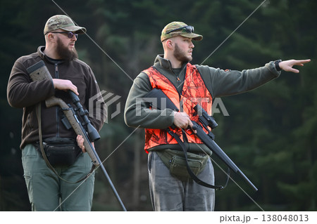 Two hunters in camo and blaze orange vest with rifles pointing and scouting in forest 138048013