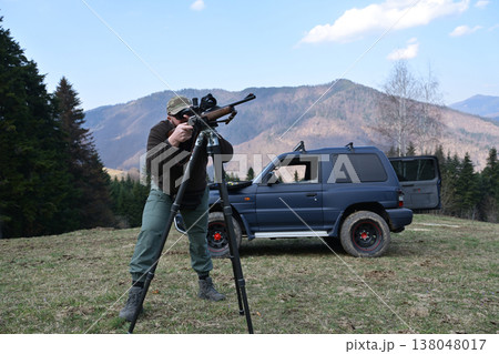 Hunter aiming a scoped rifle from tripod near SUV in mountainous forest clearing Hunter aiming a scoped rifle from tripod near SUV in mountainous forest clearing 138048017