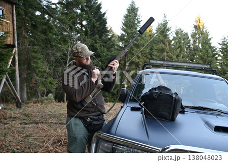 Hunter preparing a scoped rifle beside an off-road vehicle in a forested hunting camp 138048023