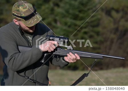 Hunter adjusting scope on a bolt-action rifle at a woodland shooting range during daytime Hunter adjusting scope on a bolt-action rifle at a woodland shooting range during daytime 138048040