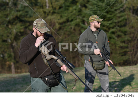 Two hunters with scoped rifles in camouflage preparing for a day of hunting in a forest clearing 138048048