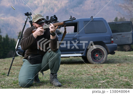 Hunter aiming scoped rifle while kneeling beside SUV during outdoor field shooting session 138048064