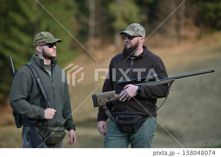 Two hunters in camouflage caps holding rifles and talking in a forested hunting field 138048074