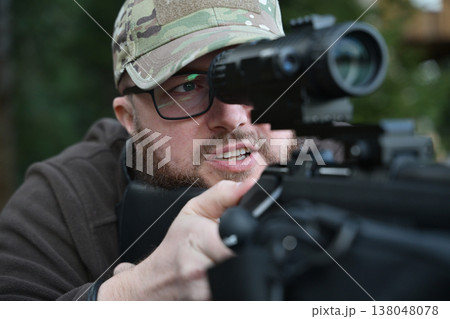 Camouflaged shooter aiming a scoped rifle outdoors with glasses and beard during tactical target practice 138048078