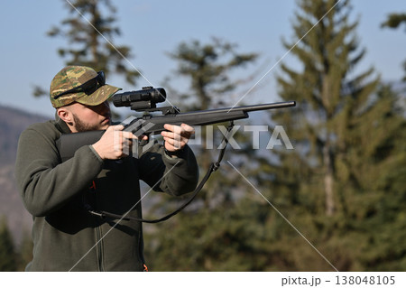 Hunter aiming rifle with scope in forest clearing wearing camouflage cap and outdoor clothing 138048105