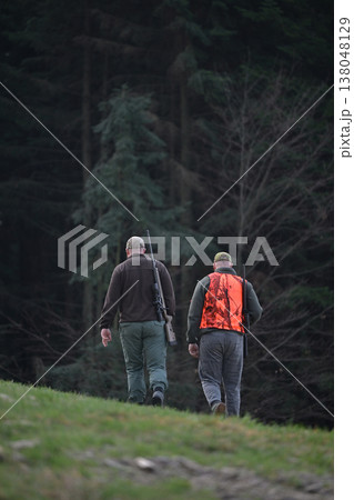 Two hunters walking into a pine forest with rifles and a bright orange safety vest 138048129
