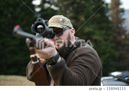 Bearded hunter aiming a scoped rifle in the woods wearing camouflage cap and outdoor jacket 138048133