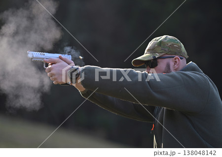 Man firing handgun at outdoor range wearing camouflage cap and sunglasses with visible muzzle smoke 138048142