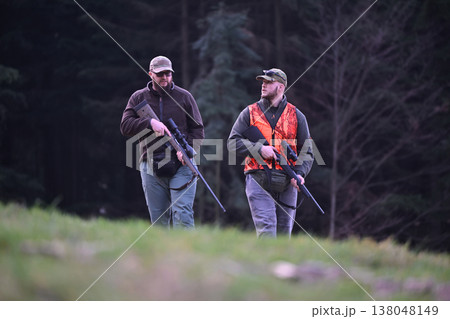 Two hunters walking through a forest field carrying rifles with one wearing a blaze orange vest 138048149