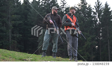 Two hunters standing with rifles in a forest wearing orange safety vest and camouflage caps 138048159