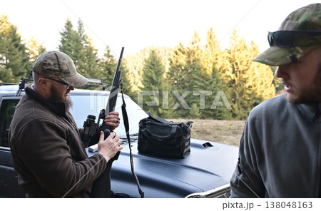 Two men preparing a rifle on a pickup truck before a hunt in a forest clearing 138048163