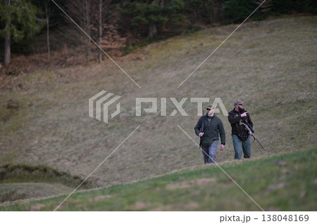 Two hunters walking with rifles on a grassy hillside near a forest during a hunting trip 138048169