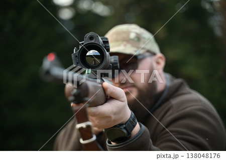 Hunter aiming a scoped rifle downrange during outdoor target practice in camouflage cap and sunglasses 138048176