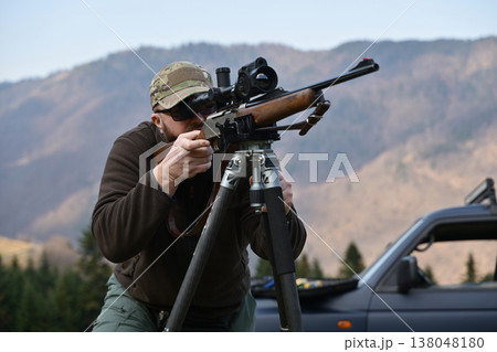 Hunter aiming a scoped hunting rifle from a tripod in mountain landscape near parked vehicle 138048180