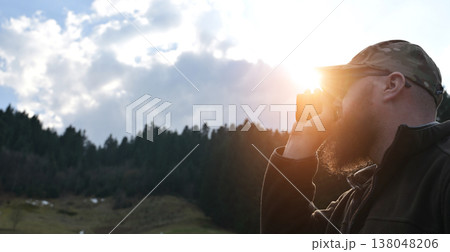 Bearded man in camo hat looking through binoculars at sunset over forested hills Bearded man in camo hat looking through binoculars at sunset over forested hills 138048206