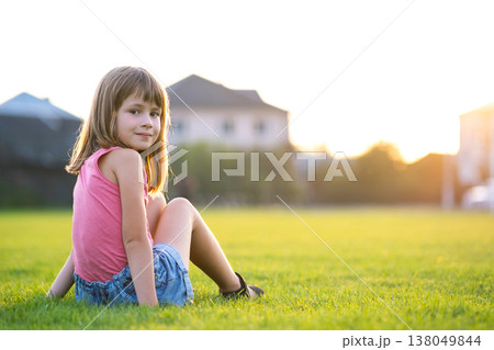 Young happy child girl resting while sitting on green grass lawn on warm summer day. Young happy child girl resting while sitting on green grass lawn on warm summer day. 138049844