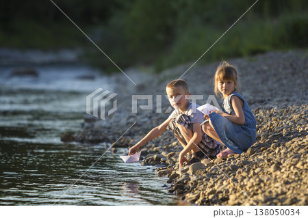 Two cute blond children, boy and girl on river bank sending in water white paper boats on bright summer blurred blue background. Joys and games of happy childhood and outdoors activities concept. 138050034