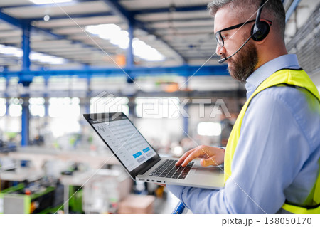 Side view of factory manager with headset and laptop standing in industrial factory. Side view of factory manager with headset and laptop standing in industrial factory. 138050170