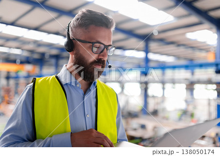 Quality manager with headset and laptop standing in industrial factory. 138050174