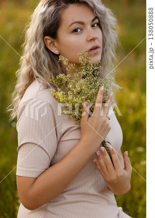 Young woman holding yellow wildflowers in meadow at golden hour, look at camera 138050888