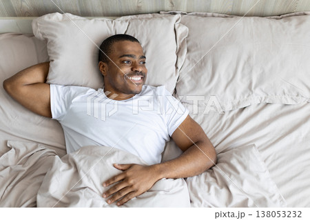 Black man lying in bed smiling and relaxed, looking away, representing comfort Black man lying in bed smiling and relaxed, looking away, representing comfort 138053232