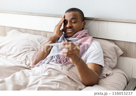 Young black man lying in bed checking temperature with thermometer Young black man lying in bed checking temperature with thermometer 138053237
